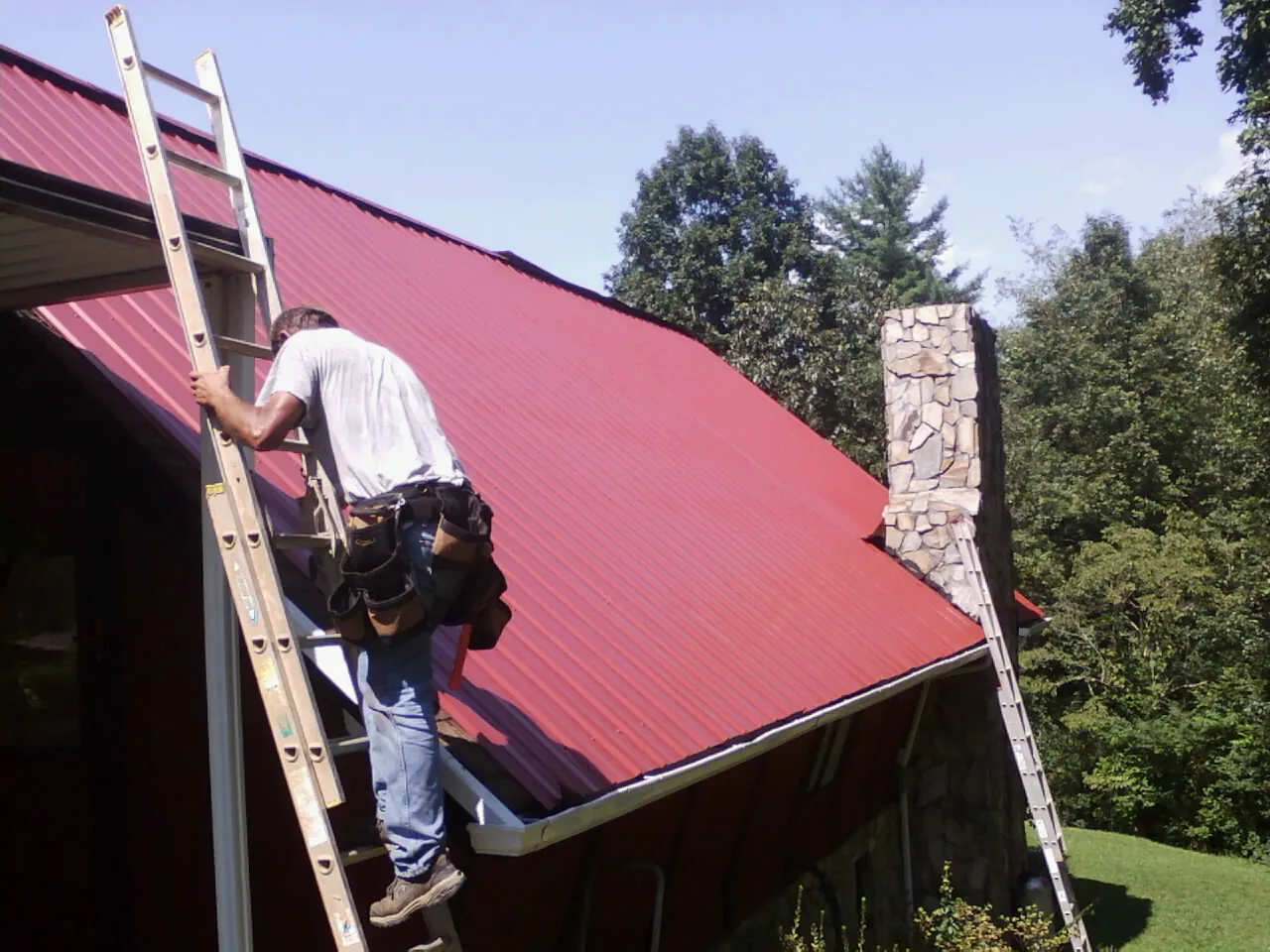 Roofer on a ladder beside a red metal roof and stone chimney — Soffit & Fascia Repair by Roofing Brothers in Cross